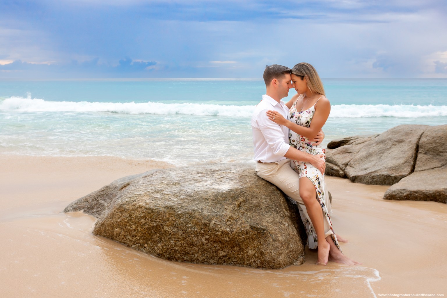 A couple sitting on a rock by the beach, embracing each other with gentle waves in the background.