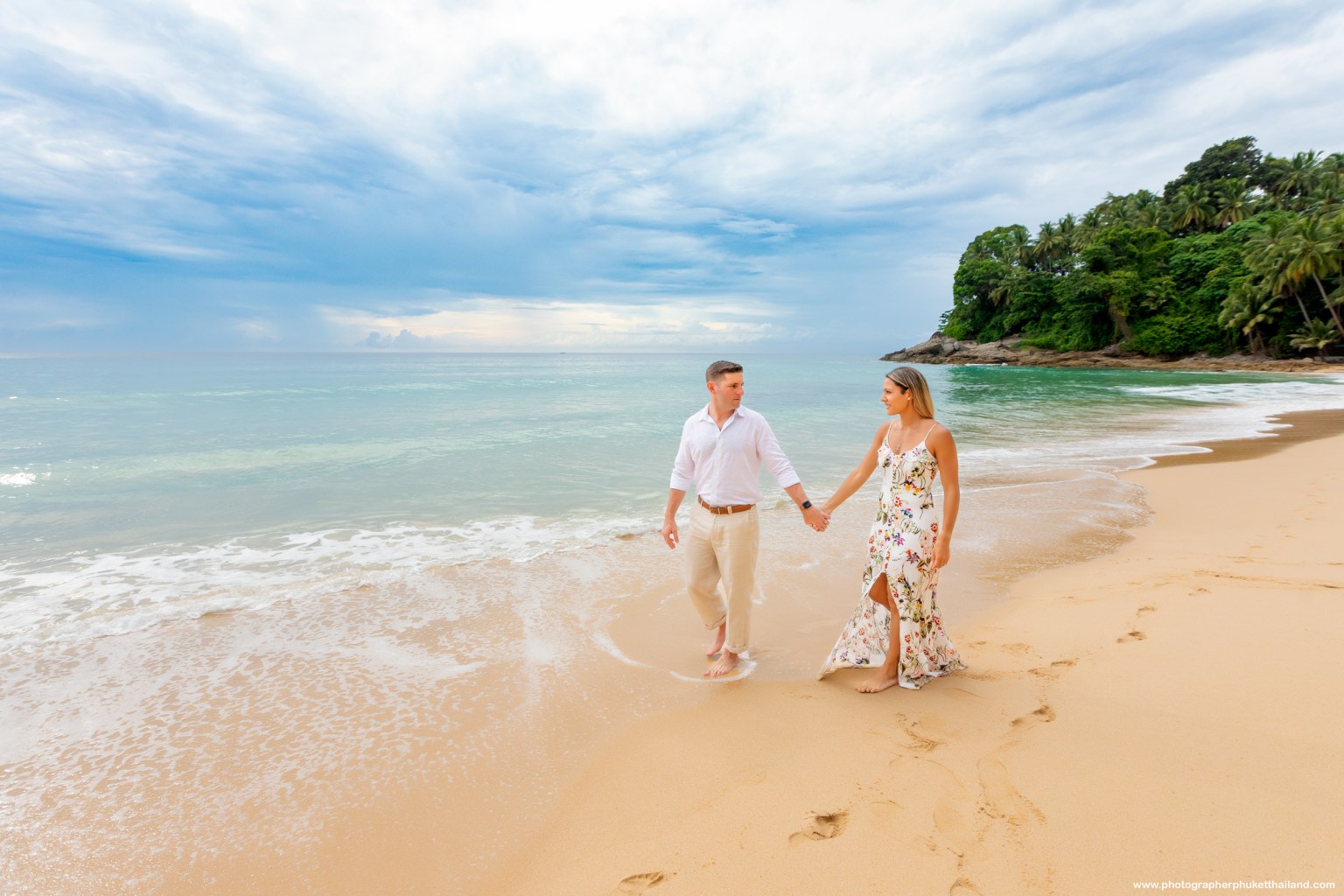 A couple walking hand in hand on a sandy beach with gentle waves and a cloudy sky in the background.