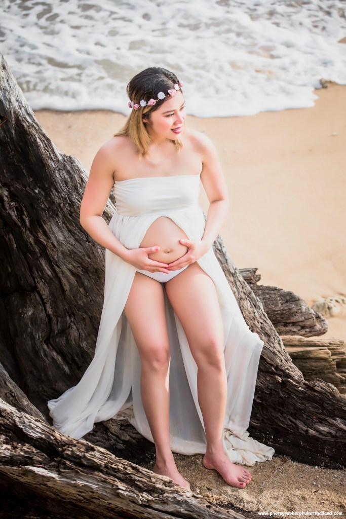 A pregnant woman wearing a flowing white dress sits on a sandy beach, holding her belly with both hands. She has a floral headband and is surrounded by driftwood, with ocean waves in the background.