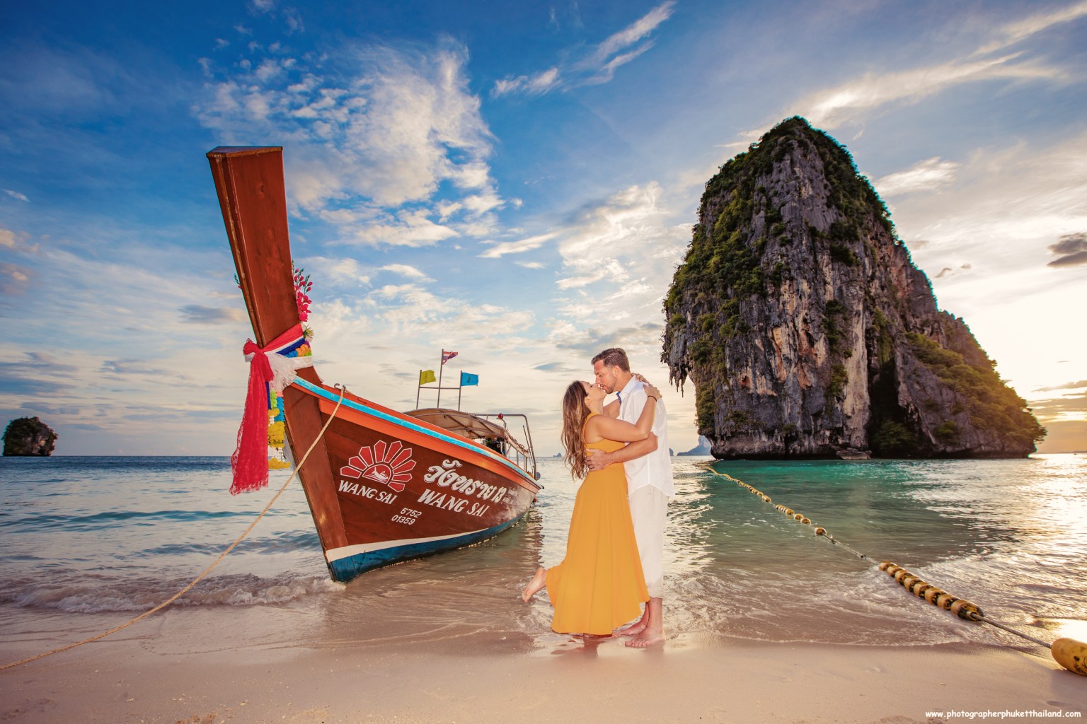 A couple embracing on a beach at sunset, with a traditional longtail boat and a limestone cliff in the background at phra nang beach railay krabi thailand