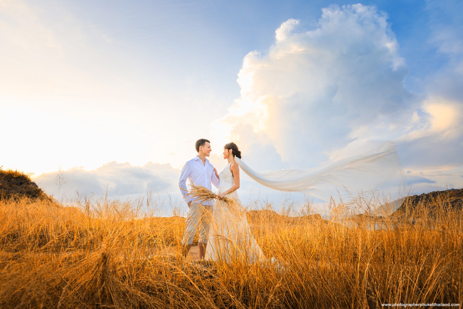 A couple stands in a field of tall grass, smiling at each other, with a soft breeze flowing through their garments and dramatic clouds in the background.