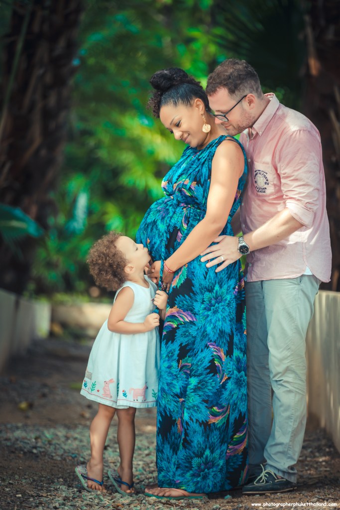 A pregnant woman in a colorful dress interacts affectionately with her husband and daughter in a lush outdoor setting.