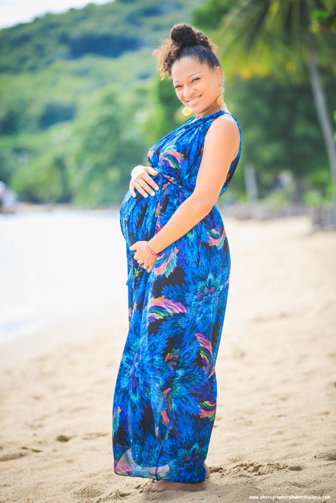Pregnant woman smiling while standing on the beach, wearing a flowing blue floral dress.