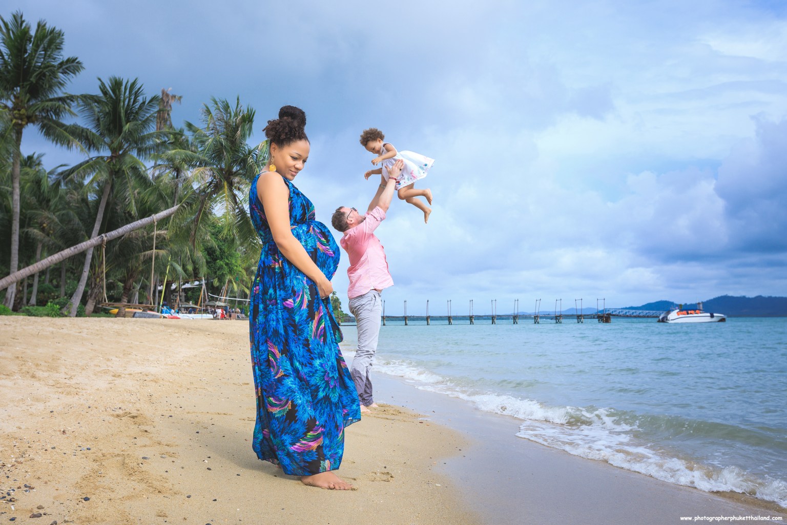 A pregnant woman in a colorful dress stands on the beach while a man lifts a young girl in the air, with palm trees and a pier in the background.
