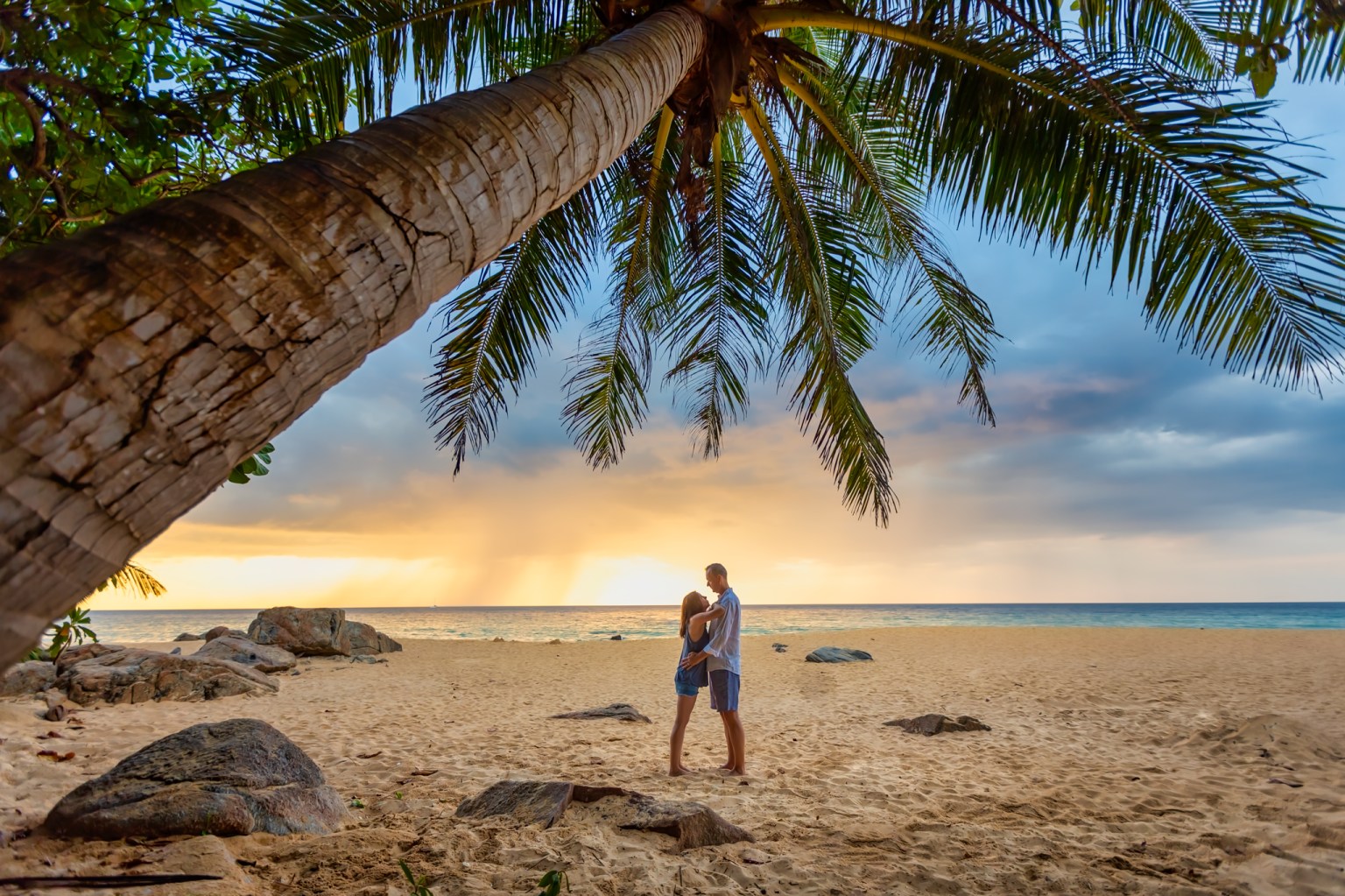 A romantic outdoor photo shoot on a beach at sunset, featuring a couple embracing under a palm tree, surrounded by rocks and sandy beach.