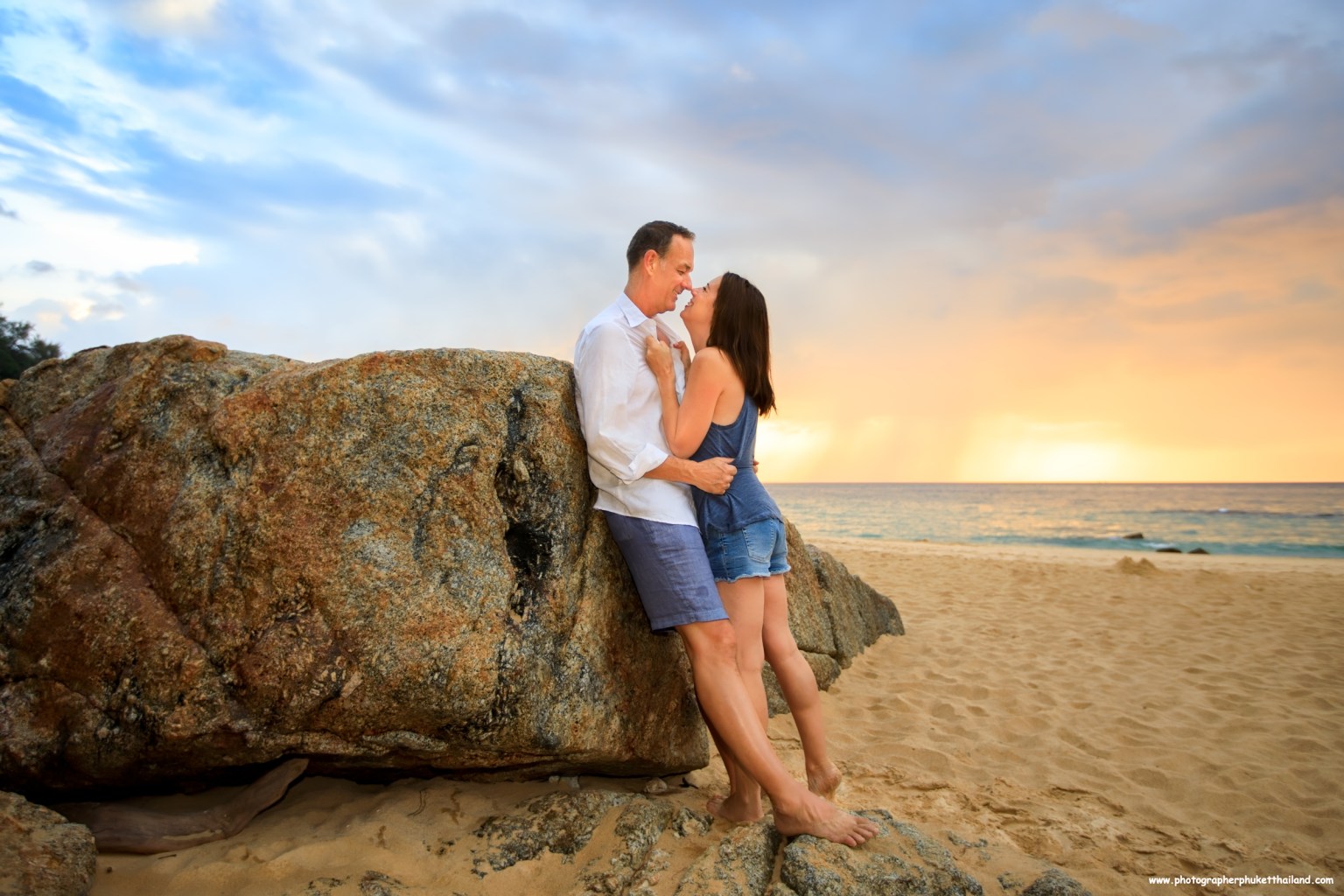 A couple embracing on the beach near a large rock as the sun sets, creating a romantic atmosphere with a colorful sky.