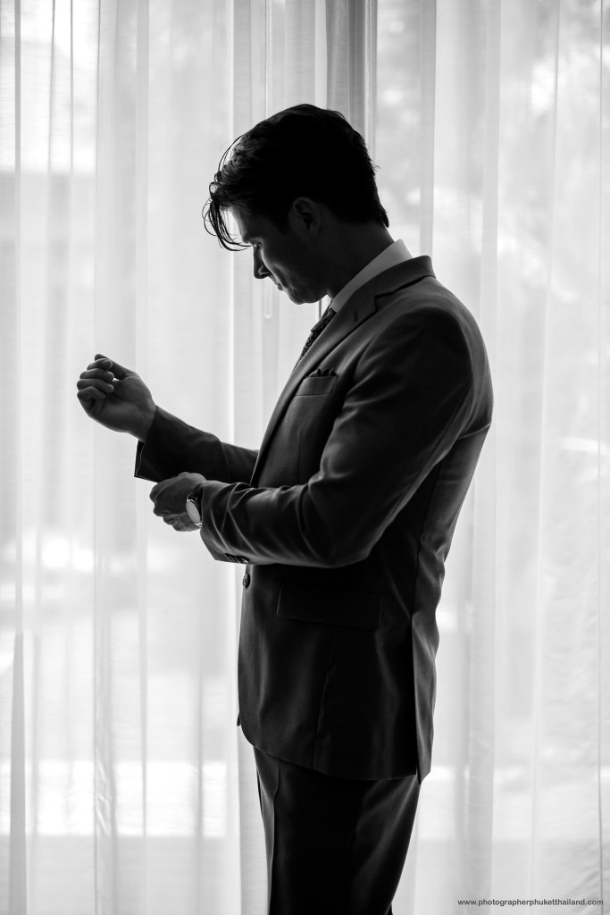 A groom adjusting his cufflinks while looking down, standing in front of sheer curtains, captured in black and white.