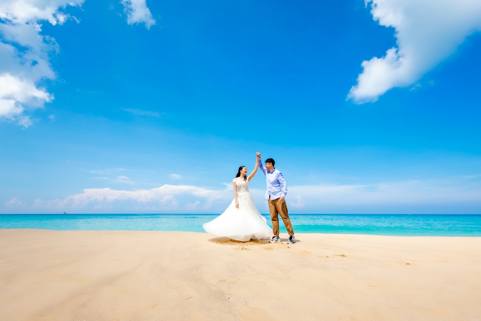A couple joyfully dancing on a sandy beach with a clear blue sky and ocean in the background.