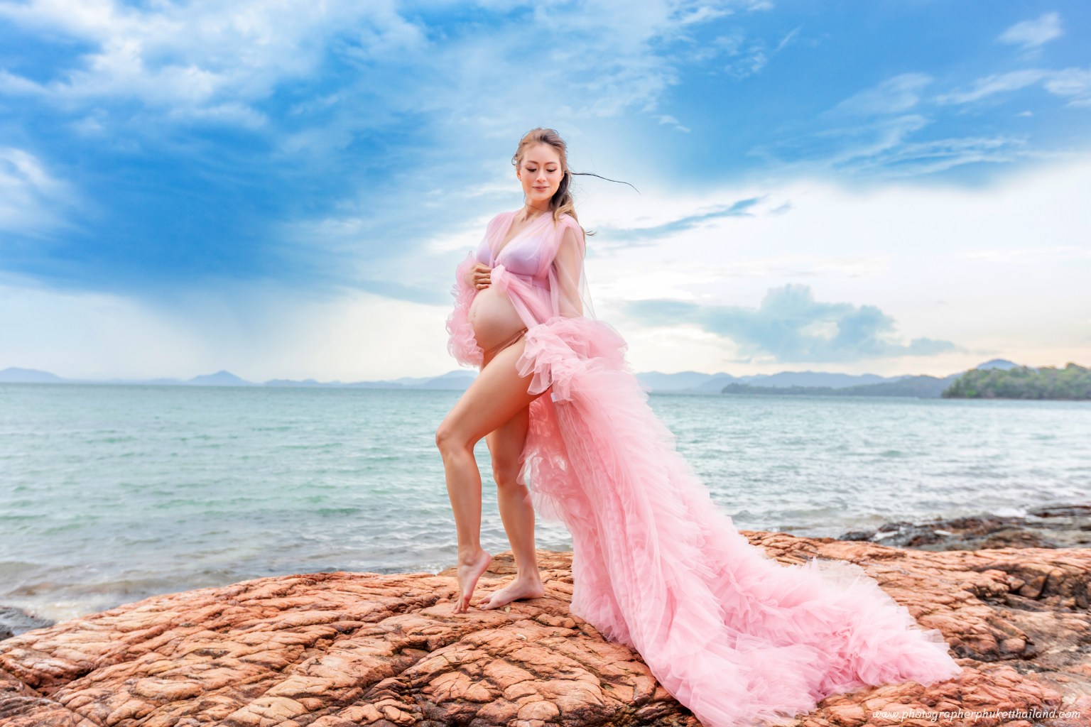 A pregnant woman wearing a pink flowing dress stands gracefully on a rocky shoreline with the ocean and sky in the background.