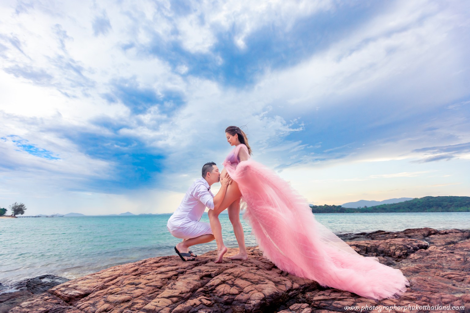 A couple enjoys a romantic moment on a rocky beach, with the woman wearing a flowing pink dress and the man kneeling beside her, set against a backdrop of blue skies and water.