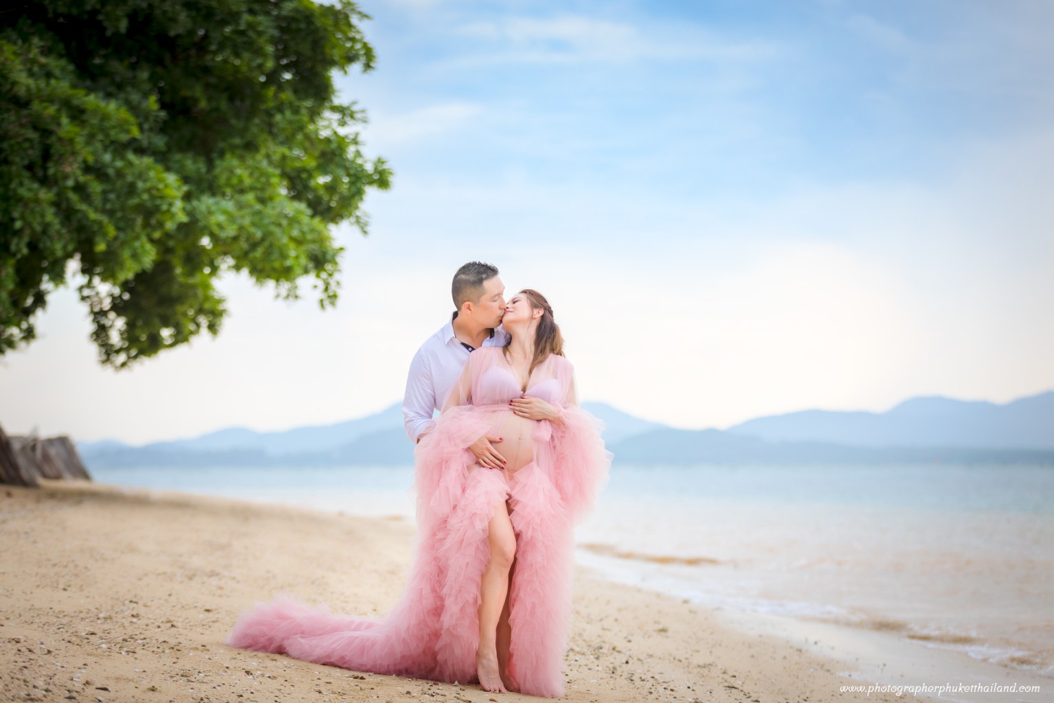A couple sharing a kiss on the beach, the woman wearing a flowing pink outfit, with gentle waves and mountains in the background.