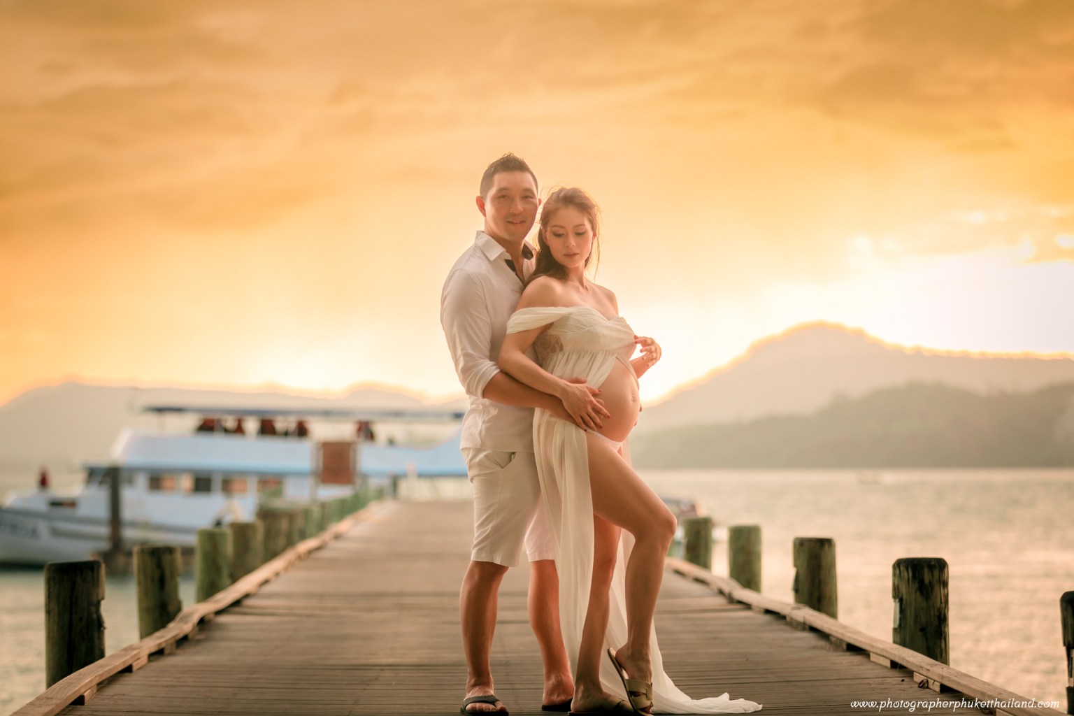 A couple poses lovingly on a wooden pier at sunset, with a soft orange glow illuminating the scene, the woman in a flowy white dress highlighting her baby bump.