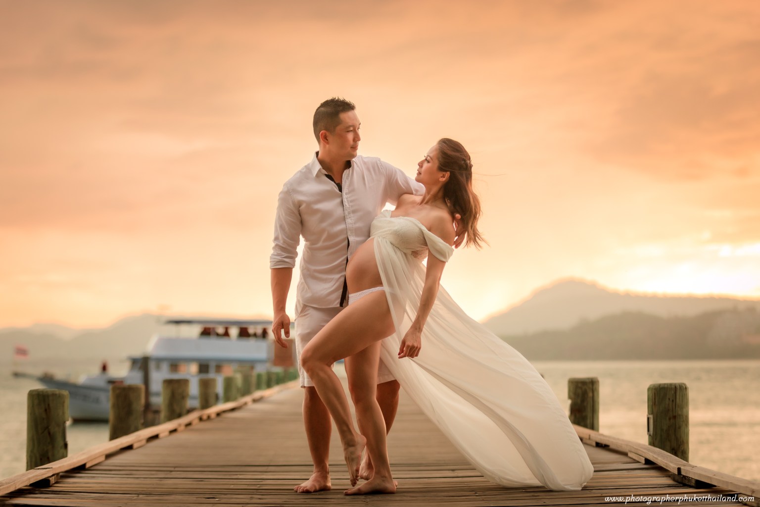 A couple posing on a wooden pier during sunset, with the woman in a flowing white dress and the man in a white shirt, showcasing a romantic moment.