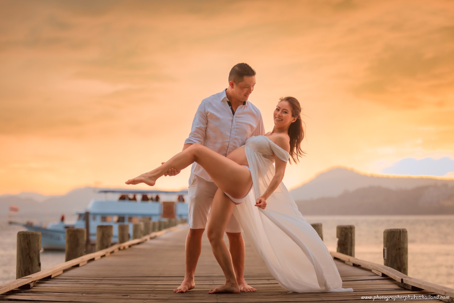 A couple posing on a wooden pier during sunset, with the woman in a flowing white dress and the man in a light shirt and shorts.