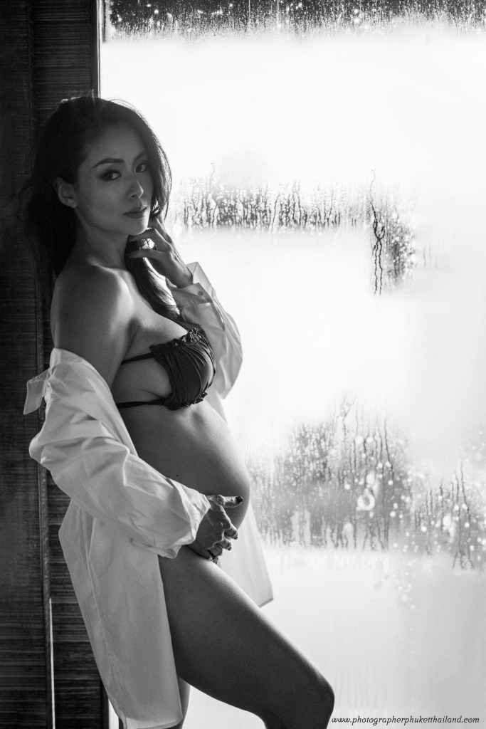 A pregnant woman in a black bikini and oversized white shirt poses in front of a fogged glass window, creating a soft and intimate atmosphere.