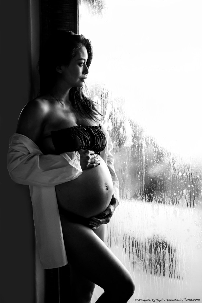 A pregnant woman wearing a black strapless top and a white shirt, standing by a fogged glass window, looking thoughtfully outwards in a black and white photograph.