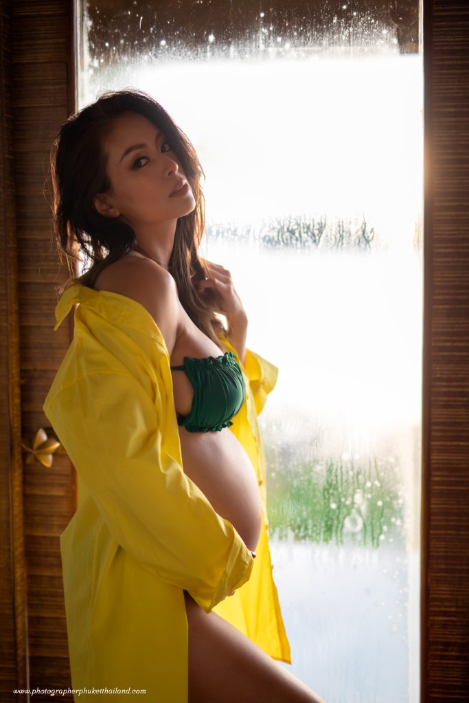 A pregnant woman in a green bikini top and a yellow shirt poses near a window with condensation, highlighting her silhouette against a bright background.
