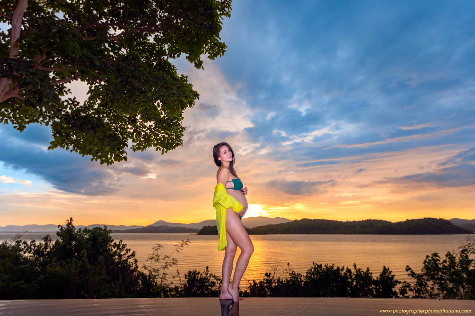 A pregnant woman poses confidently in a bikini with a yellow cover-up, standing near a waterfront during sunset. The sky is filled with a mix of clouds and vibrant colors, creating a serene and picturesque backdrop.