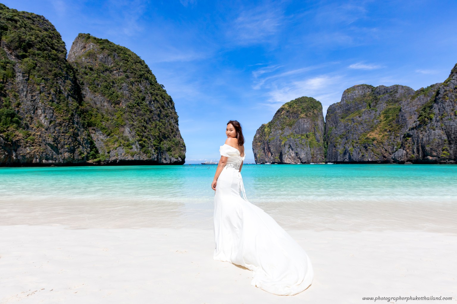 A woman in a white dress stands on the beach at Maya Bay, surrounded by stunning limestone cliffs and clear turquoise water.