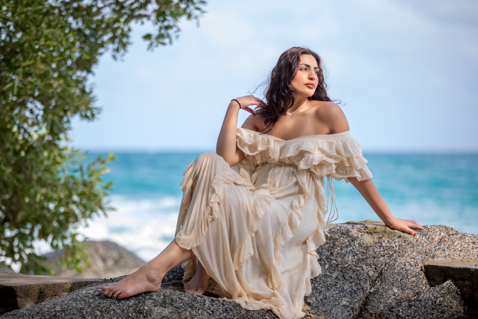 A woman in a flowing cream dress poses on a rock by the ocean, with turquoise waves and green foliage in the background.