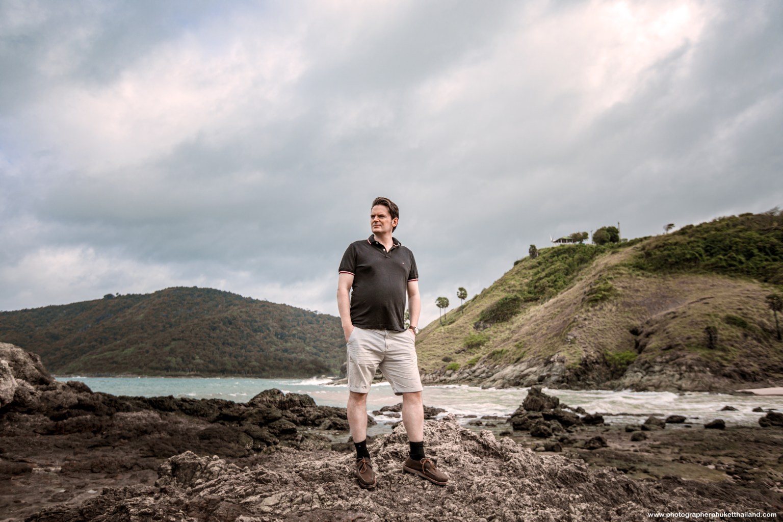 A man standing on rocky terrain by the beach, wearing a black polo shirt and shorts, with hills in the background under a cloudy sky.