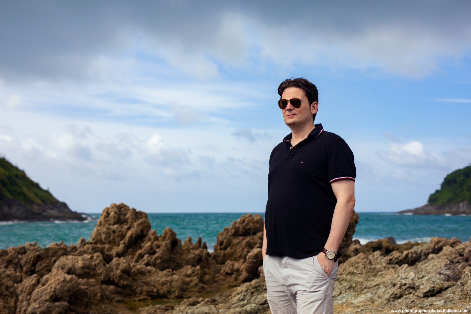 A person in sunglasses stands on rocky terrain near the ocean with a cloudy sky in the background.