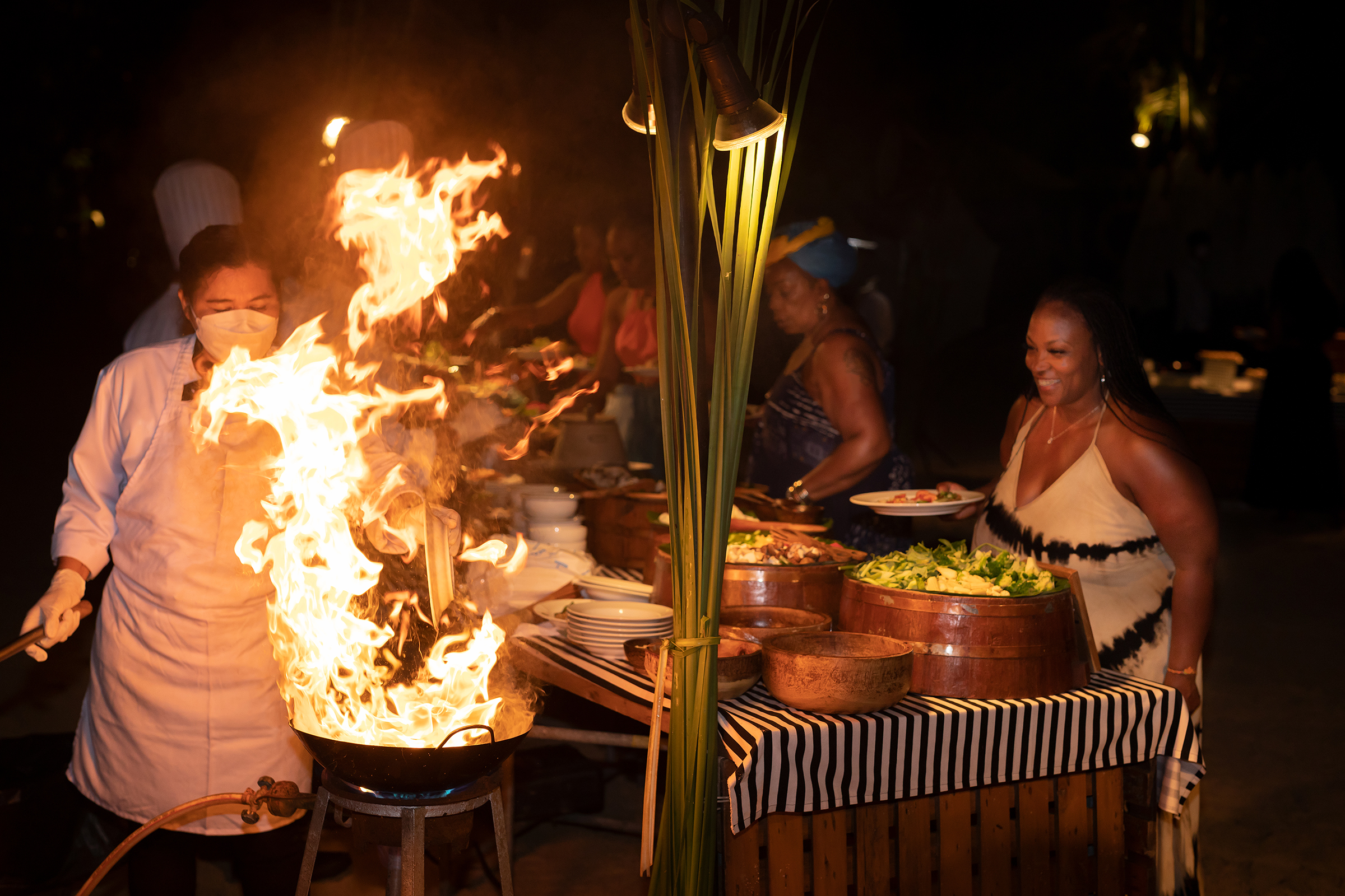 Chef preparing food with flames at a beachfront event, surrounded by guests enjoying a buffet.