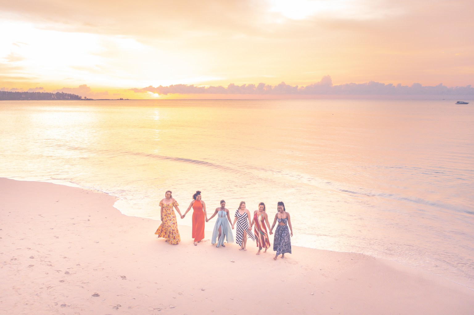 A group of seven women holding hands and walking along a beach during sunset, with colorful dresses and a calm sea in the background.