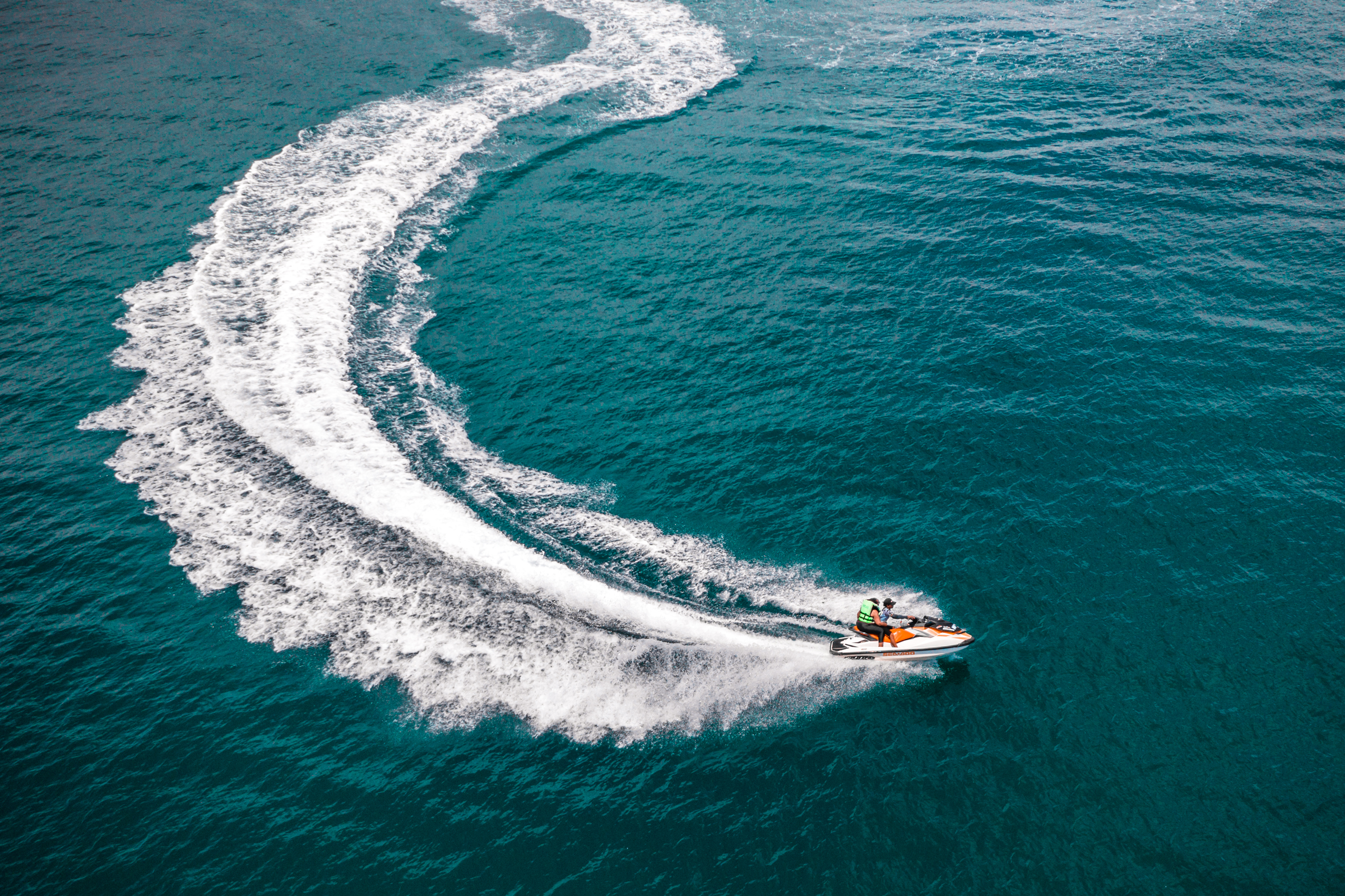 Two people riding a jet ski on turquoise water, creating white wake patterns behind them.