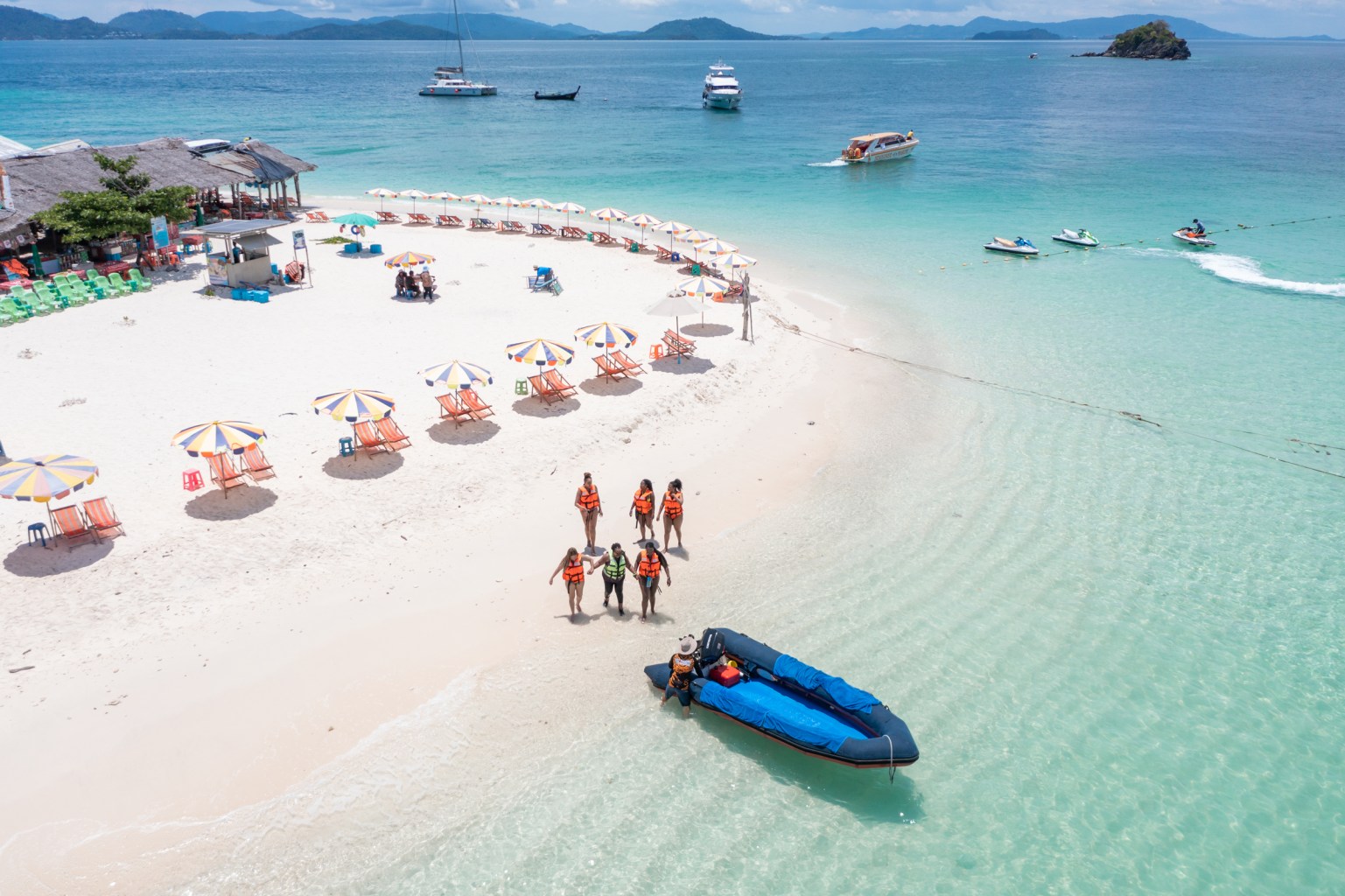 Aerial view of a beach with colorful umbrellas and lounge chairs, where a group of people in life jackets are approaching a boat on the shore.