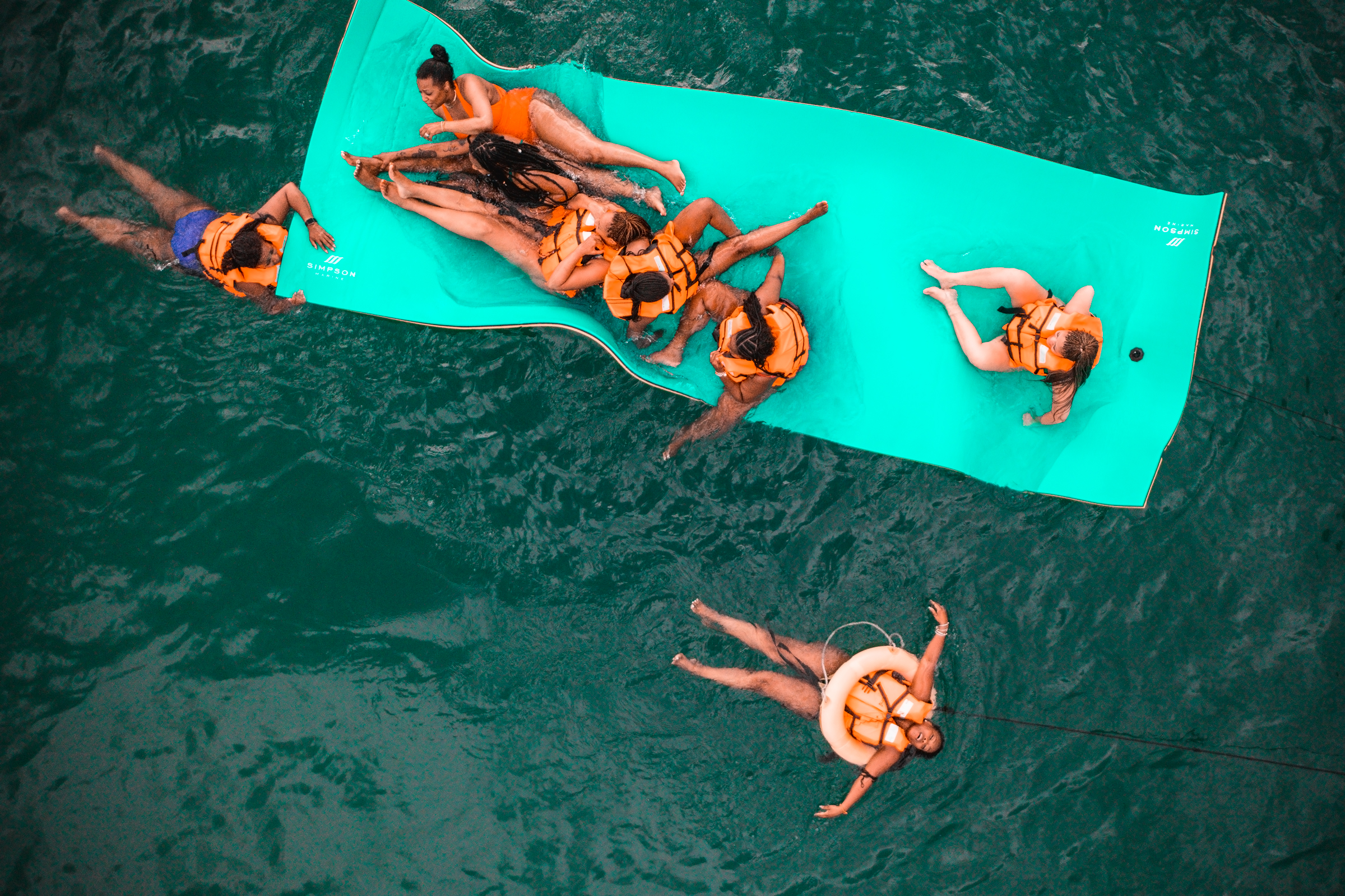 Aerial view of several people wearing life vests enjoying a green floating mat in a body of water.