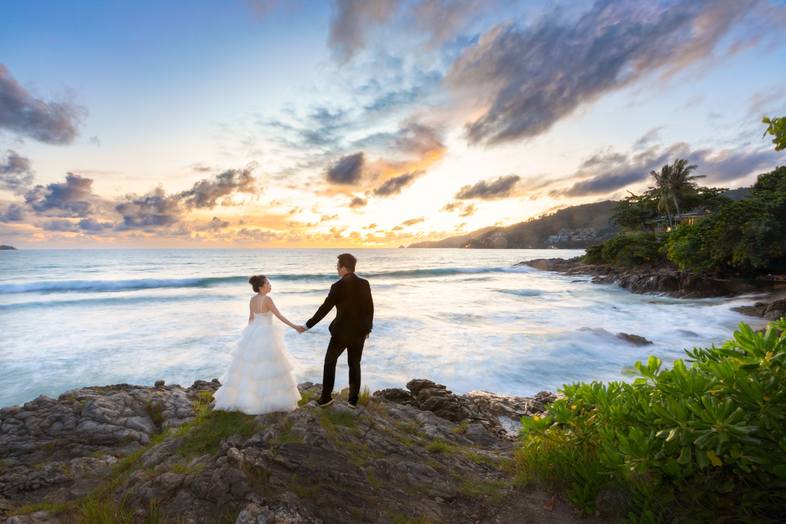 A couple in formal attire standing on rocks by the ocean at sunset, holding hands with waves gently crashing in the background.