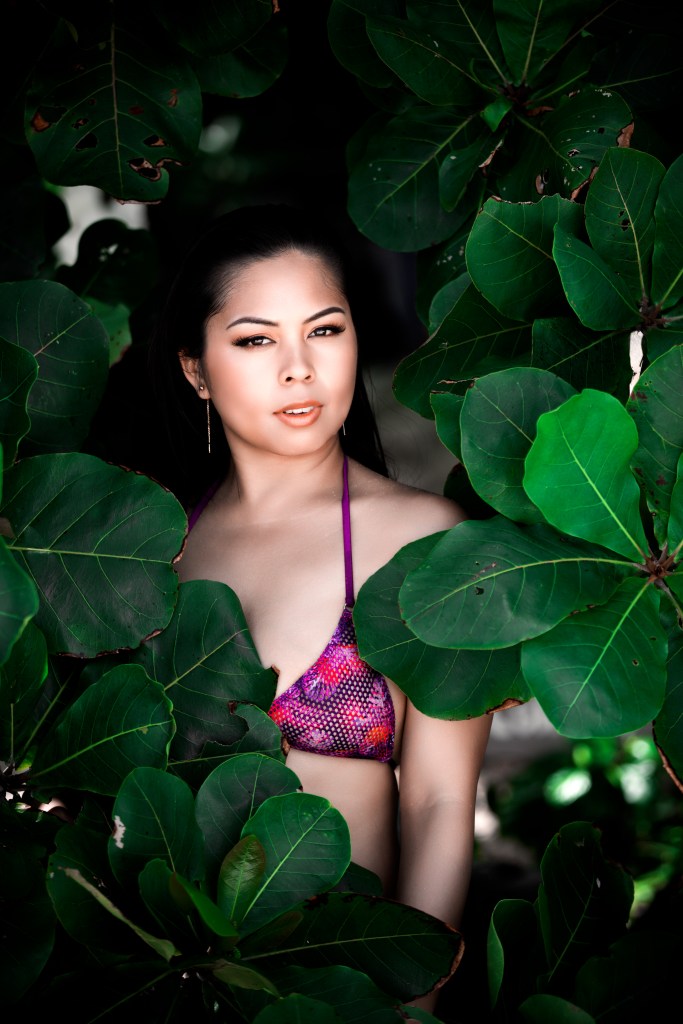 A woman in a purple bikini poses amidst large green leaves, showcasing a natural and vibrant outdoor setting.