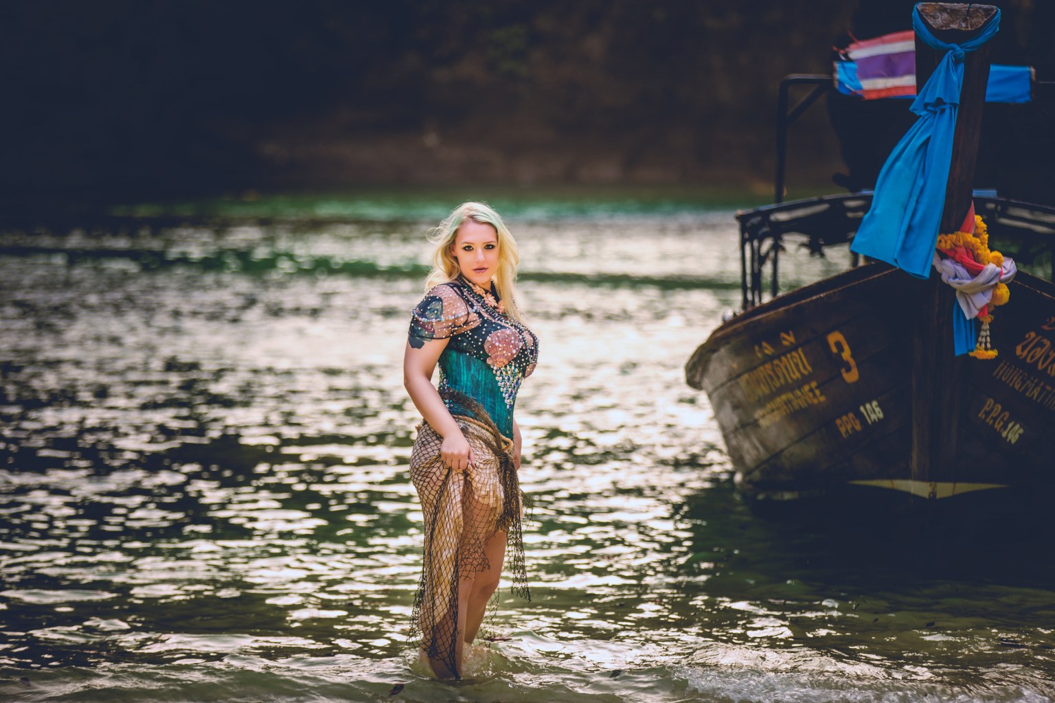 A woman wading in shallow water near a boat, wearing a sparkling dress and a fishnet cover-up.