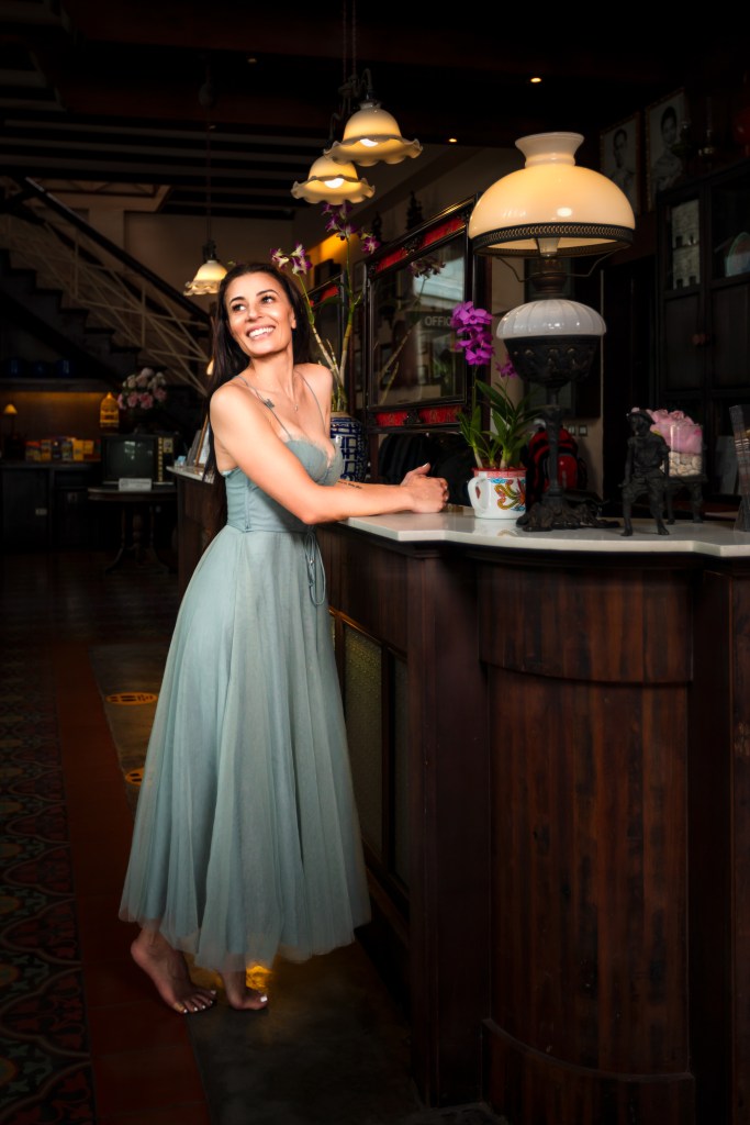 A smiling woman in a light blue dress stands at a bar counter, surrounded by decorative lamps and flowers, in a cozy indoor setting.