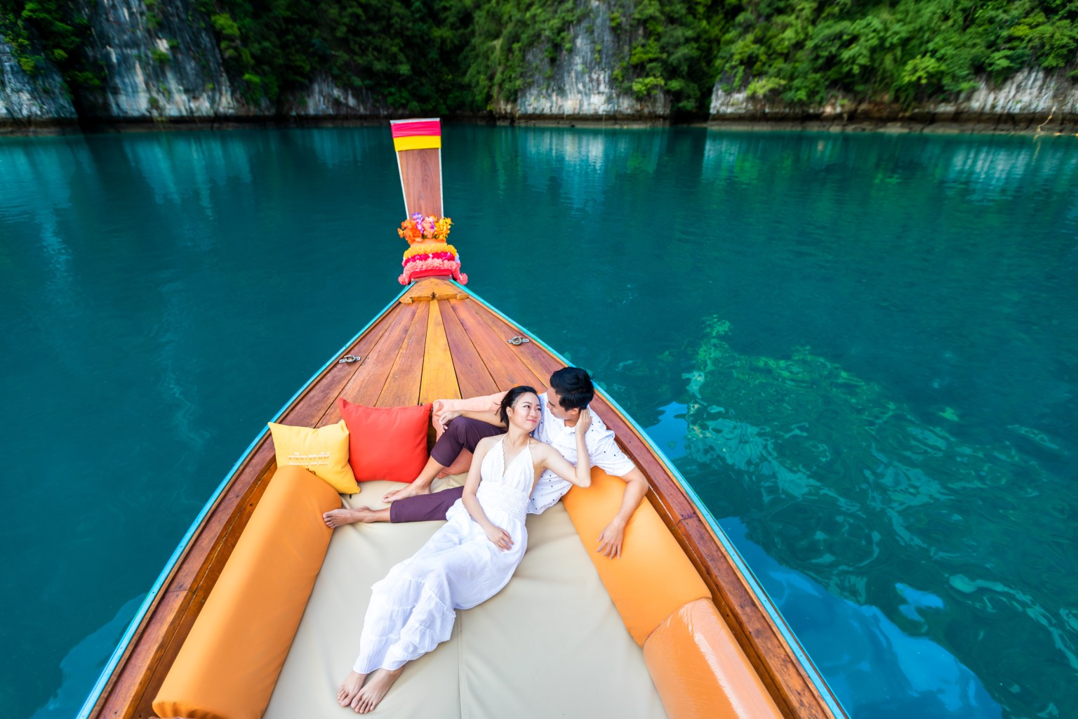 A couple relaxing on a long tail boat at Pileh Lagoon, Phi Phi Island, Krabi, surrounded by emerald water and lush greenery.