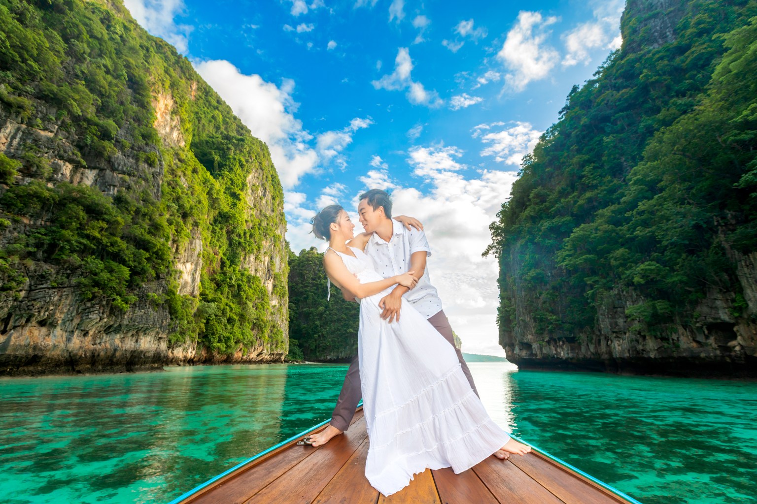 A couple embracing each other on a boat surrounded by lush green cliffs and clear turquoise water under a blue sky with clouds.