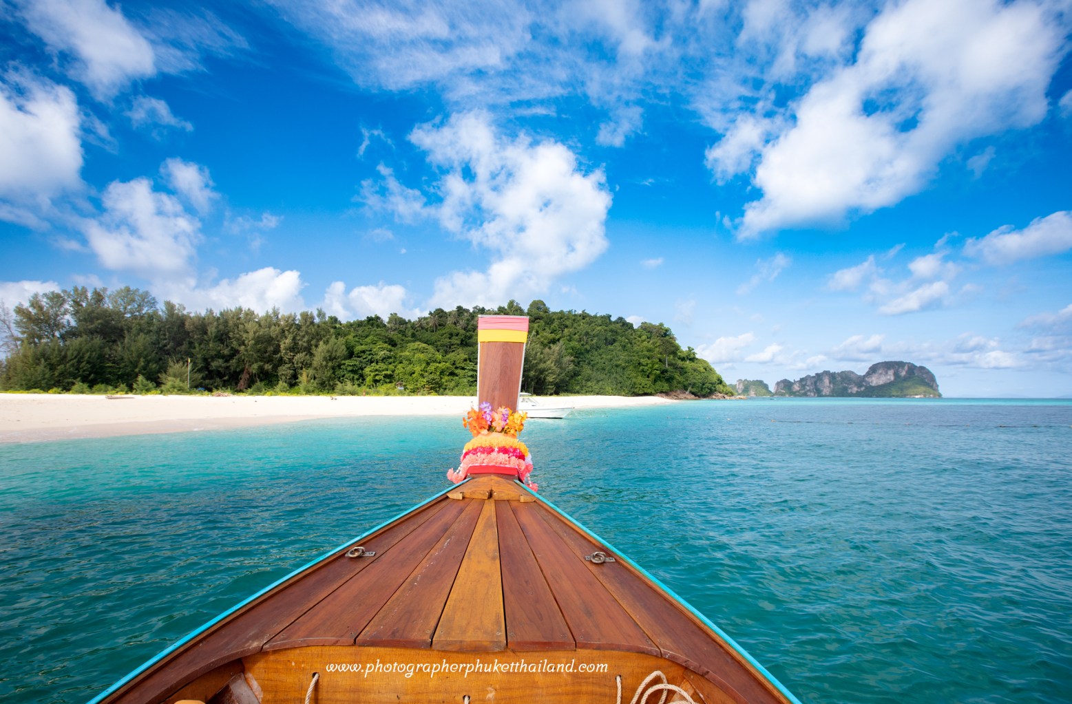 View from the front of a traditional Thai long-tail boat on turquoise water, approaching a sandy beach surrounded by greenery and mountains under a blue sky with fluffy clouds at bamboo island phi phi krabi..