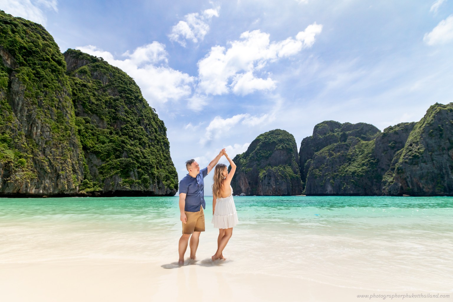 A couple holding hands and dancing on the beach at Maya Bay, surrounded by green limestone cliffs and clear turquoise water under a blue sky.
