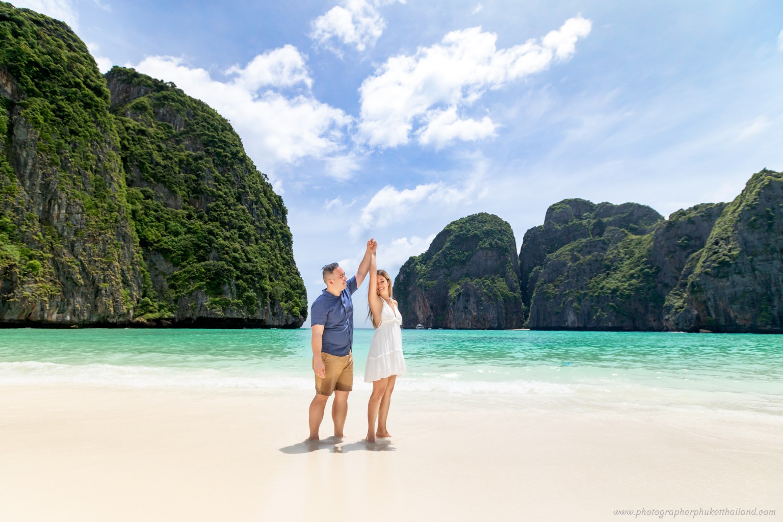 Couple holding hands on a sandy beach at Maya Bay, surrounded by lush limestone cliffs and clear turquoise water.