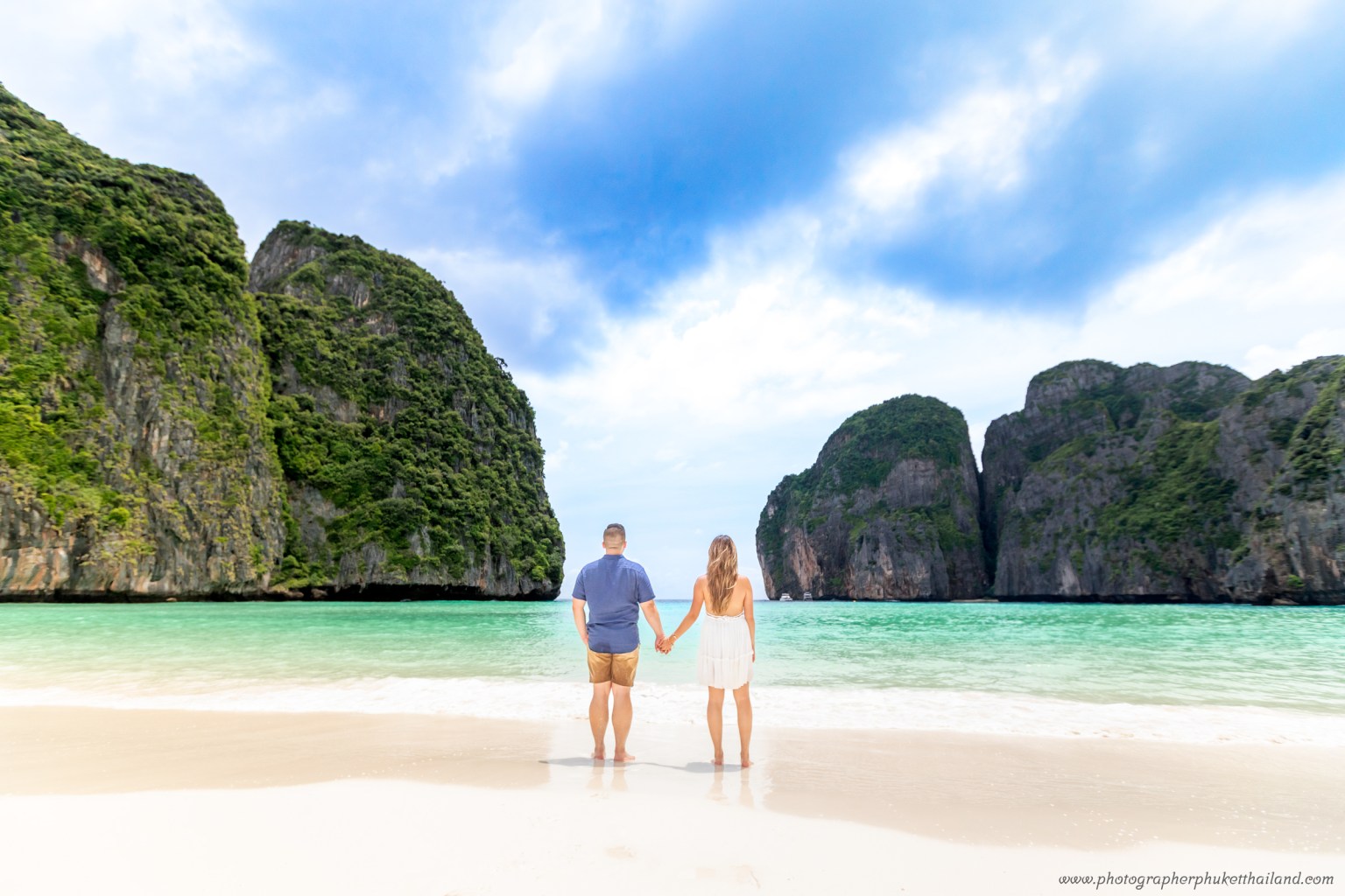 A couple standing hand in hand on the beach at Maya Bay, with limestone cliffs and turquoise water in the background.