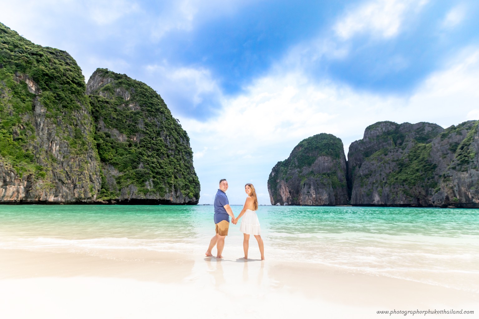 A couple holding hands on the beach at Maya Bay, surrounded by lush limestone cliffs and clear turquoise water.