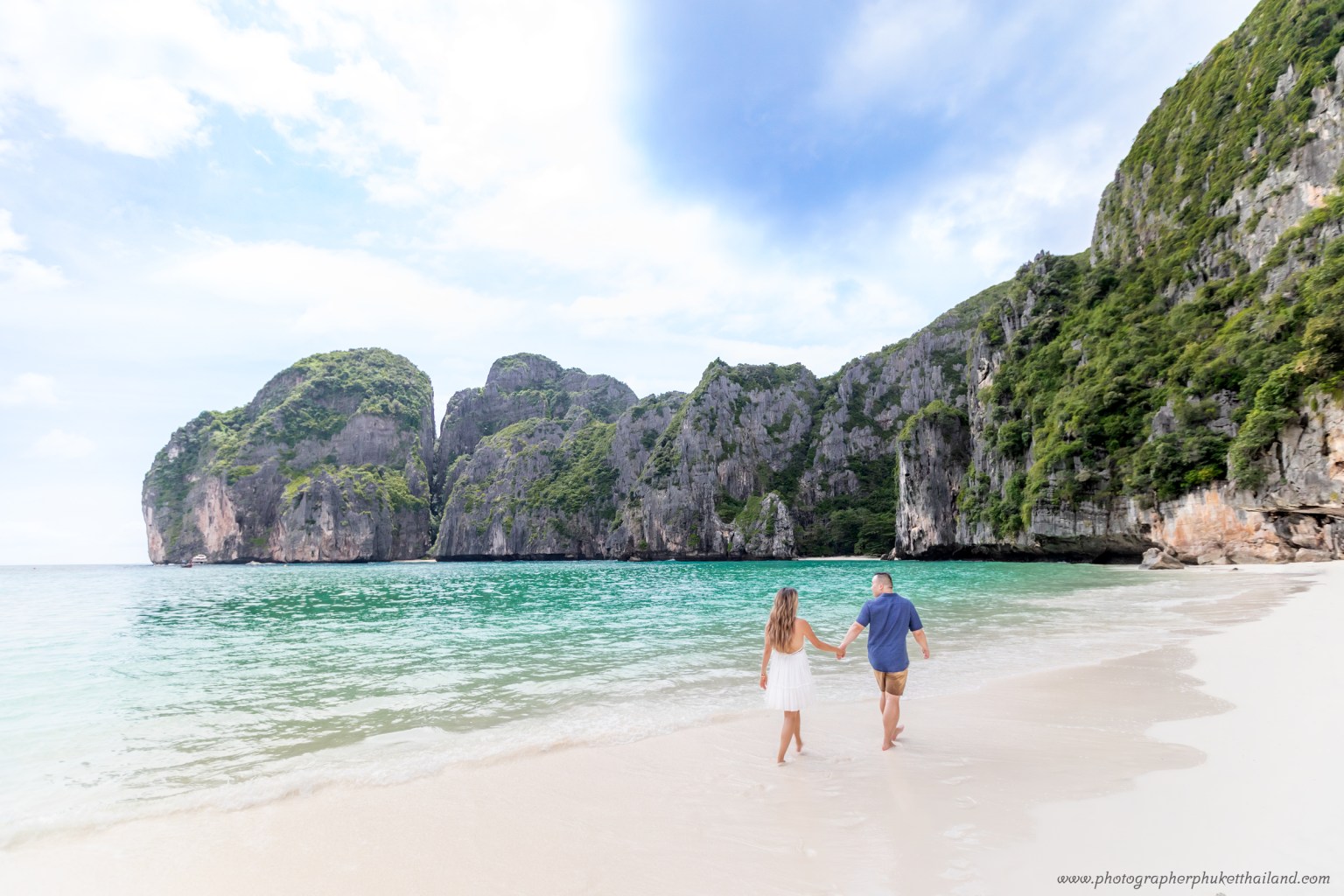 A couple walking hand in hand along the sandy beach of Maya Bay, surrounded by lush limestone cliffs and clear turquoise waters.