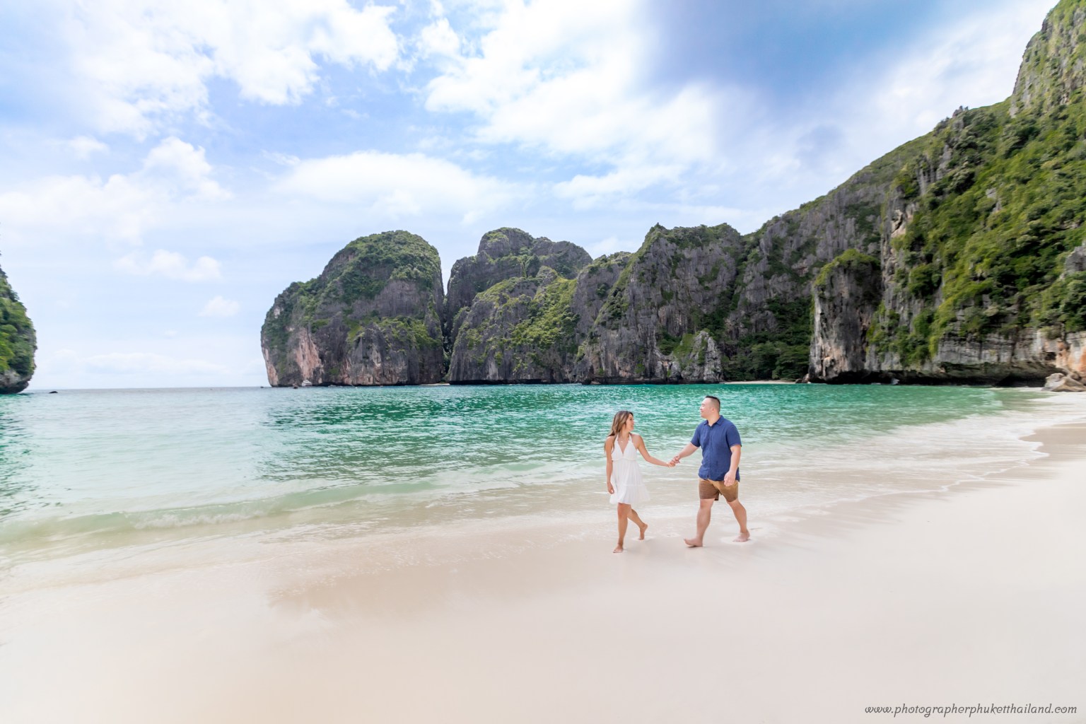 A couple walks hand in hand along the sandy beach of Maya Bay, surrounded by lush green cliffs and crystal clear waters under a partly cloudy sky.