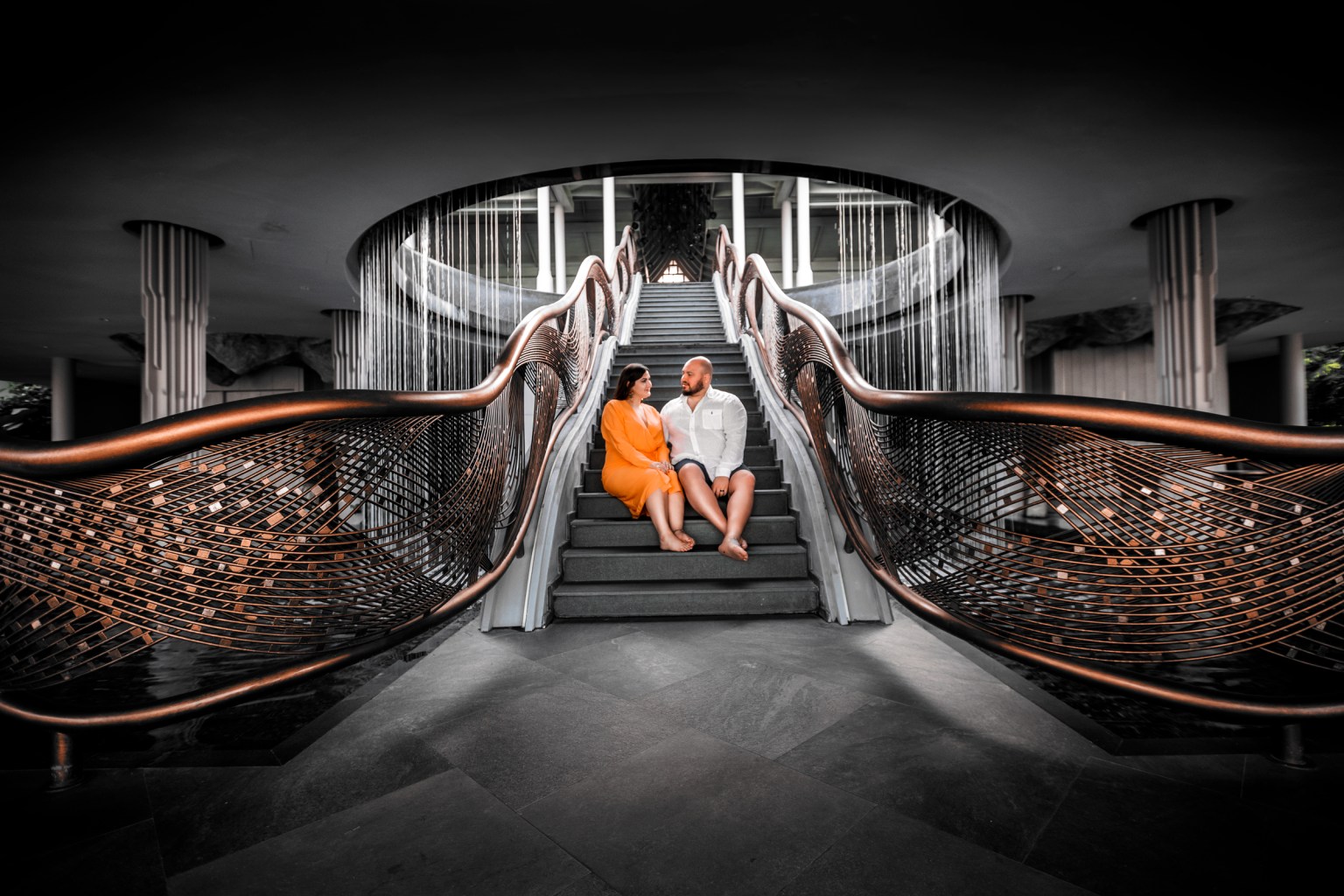 A couple sitting on the stairs of a modern hotel with a decorative railing, surrounded by a stylish interior design.