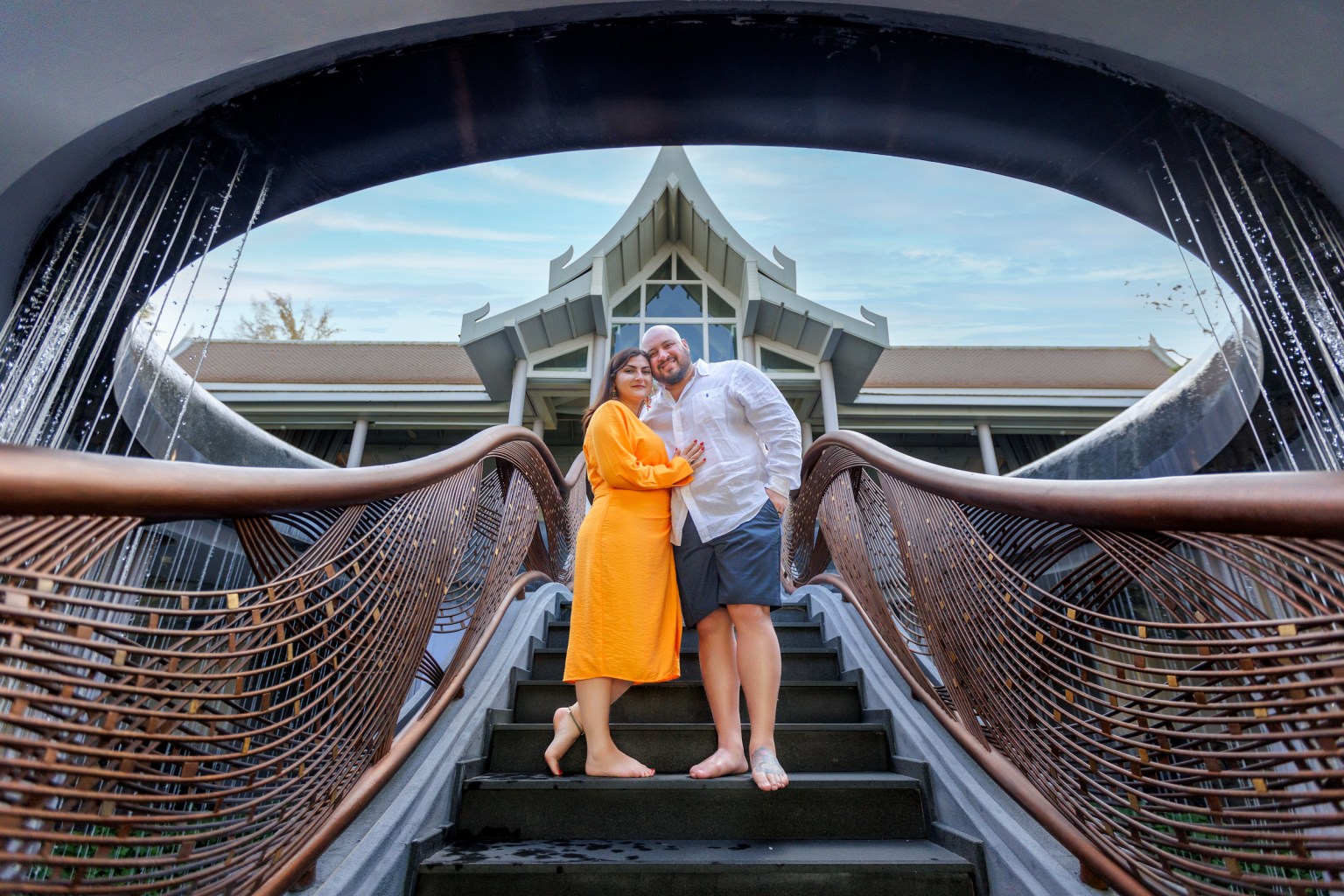 A couple poses together on a staircase with a modern arching wooden railing, surrounded by water features, under a clear sky.