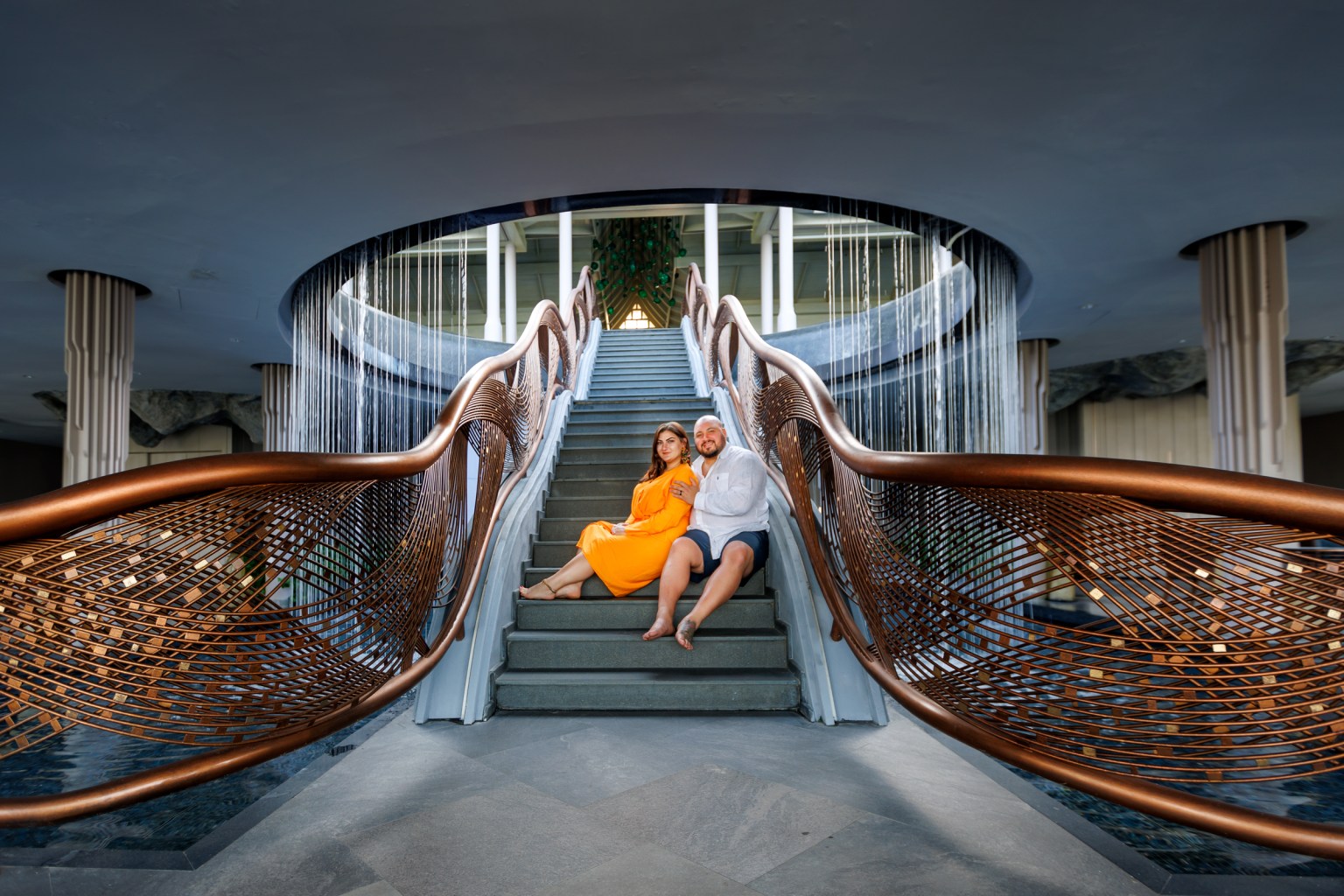 A couple seated on a stylish staircase featuring a modern copper railing, with water trickling down in the background.