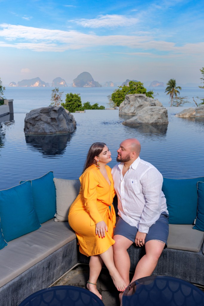A couple sitting together on a plush couch by an infinity pool overlooking the ocean, with rock formations and islands in the background under a blue sky.