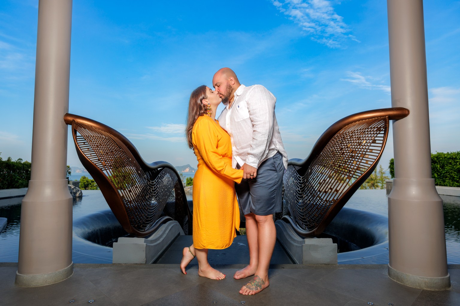 A couple kissing in a serene outdoor setting with a water feature, surrounded by modern architecture and blue skies.