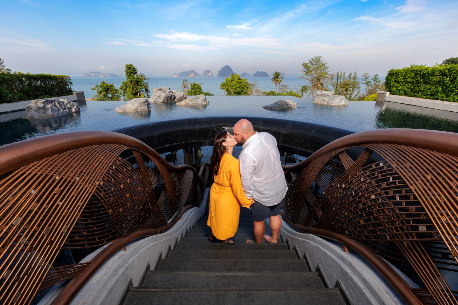A couple shares a kiss while holding hands on a stairway, overlooking a serene body of water with distant islands and green foliage in the background.